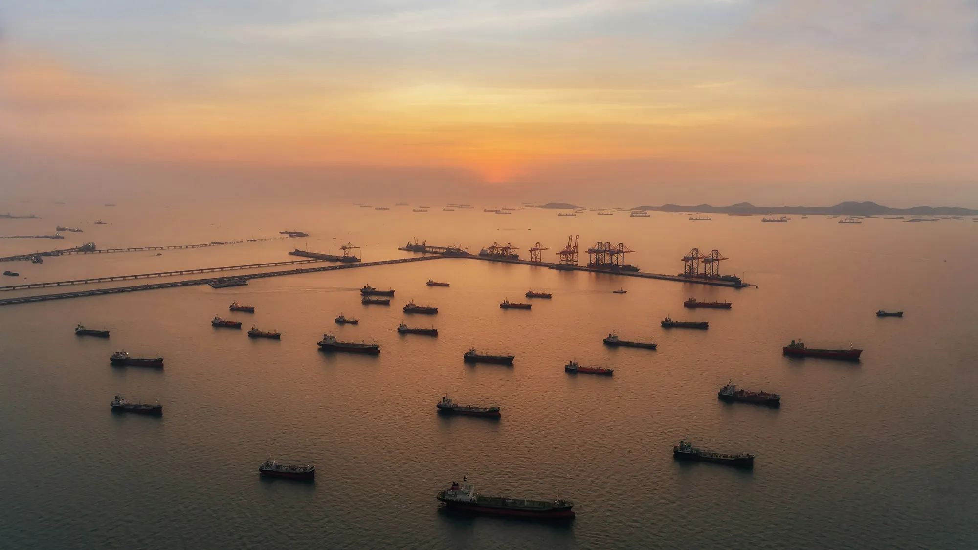 Sunset view over a harbor with numerous cargo ships anchored near industrial docks.